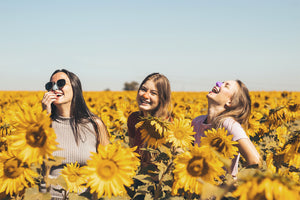 three girls laughing and smiling in a sunflower field using the colorful spf protection Nöz sunscreen for their nose that are reef safe, eco friendly and environmentally safe.